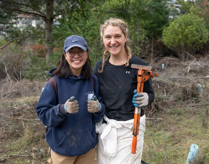 Two smiling people wearing work gloves pose in front of plants and trees. One holds a gardening tool. 