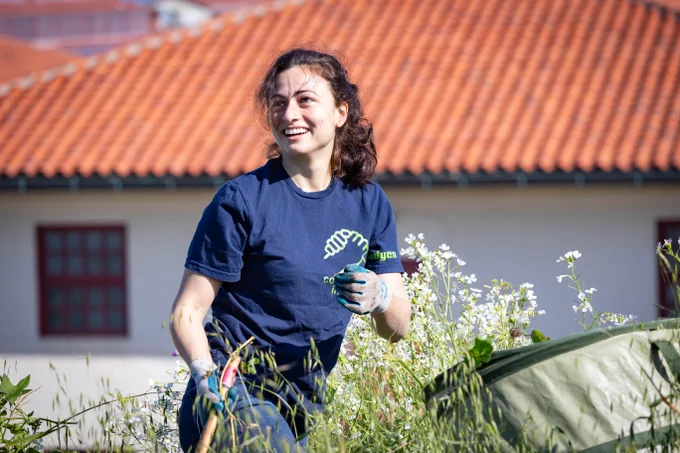 A smiling person holding a tool looks up from their work next to a large brush canister. A Fort Mason building with a red tiled roof is in the background.