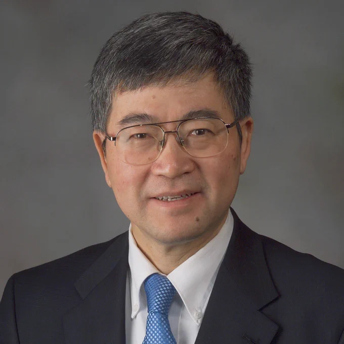 Professional portrait of Ge Wang, wearing a dark blue suit, white dress shirt, and light blue tie.