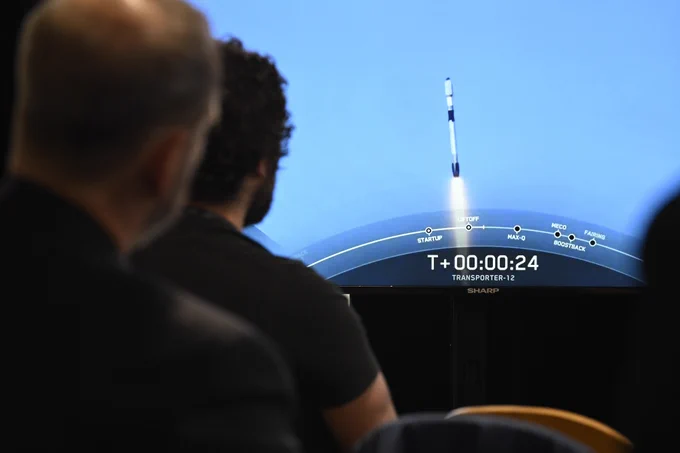 A television screen shows a rocket lifting off, while people watch from chairs.