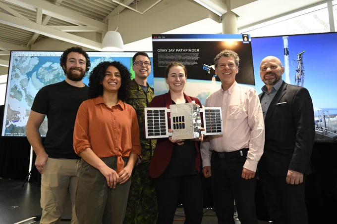 A group of smiling people stand in front of a sign that says “Gray Jay Pathfinder.” The person in the centre of the group holds a model of a satellite.