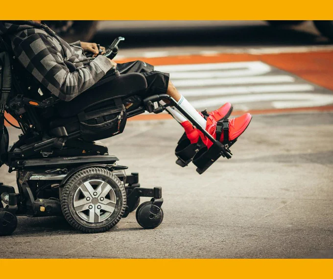 A person in a wheelchair. The person's face is not visible but they have their legs on the pedals of the chair and are wearing red shoes. 