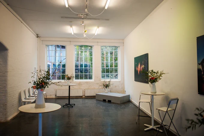 A long rectangular room with and industrial floor and three large windows on one end. Three bar tables are arranged around the room decorated with vases full of flowers. A row of chairs sits against the white walls and posters of Kaleider's artworks hang on the walls.