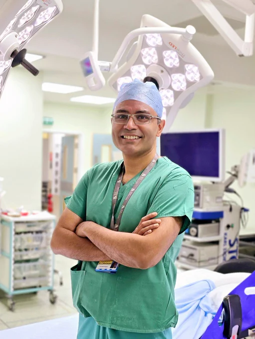 Mr Rajarshi (Rishi) Mukherjee, Consultant Surgeon in Emergency General and Major, wearing his green scrubs and surgical cap, smiling with his arms crossed in theatre in front of a surgical bed.