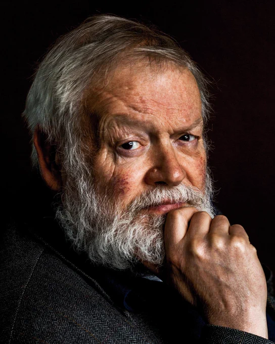 Photographic portrait of Michael Longley by Bobbie Hanvey. A person with grey hair raises their hand to their face, with a finger resting under their lip. They look directly at the camera. They wear a dark jacket and the background is dark.