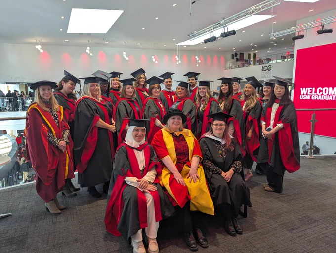 A group of graduates and faculty members posing for a photo indoors at a graduation ceremony. 