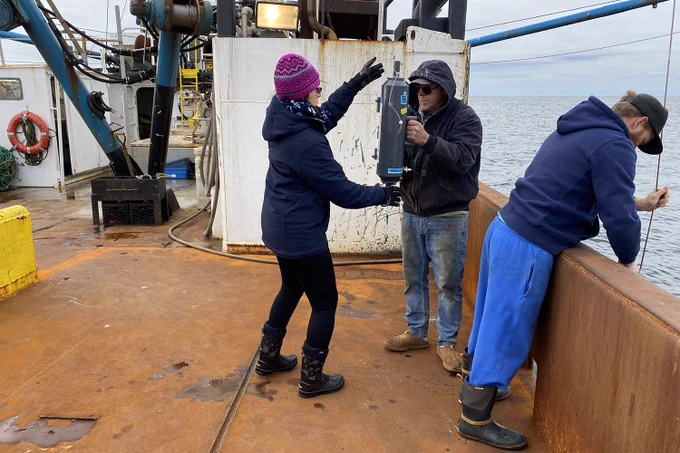Three people work on the deck of a fishing vessel. A woman on the left, with a pink winter hat, is getting a sampling bottle of about 5 litres from a man (in the middle) with sun glasses and a dark blue hoodie. Another man (on the right) is holding on to a wire while looking down at the surface of the ocean. 