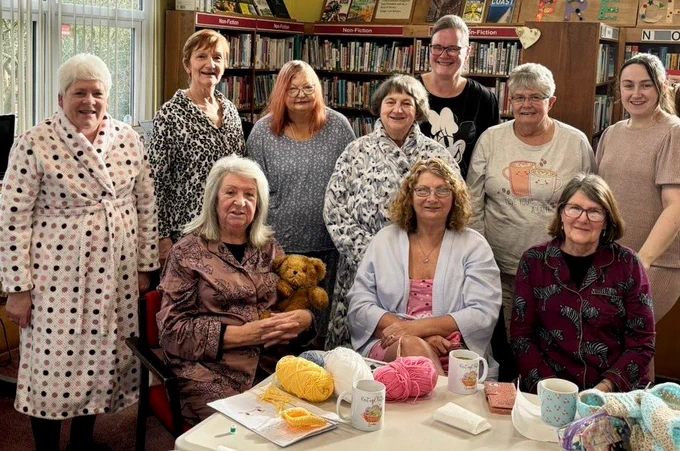 A group of women all wear pyjamas. In front of them on a table there are balls of yarn.