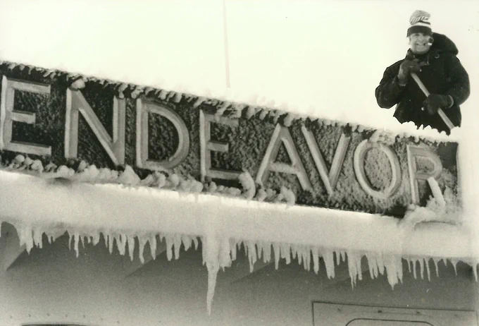 A man stands atop an ice-covered Endeavor sign, in black and white. 