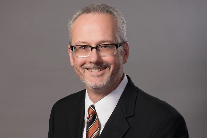 A portrait of a smiling individual wearing glasses, a dark suit, and a striped tie, set against a neutral gray background.
