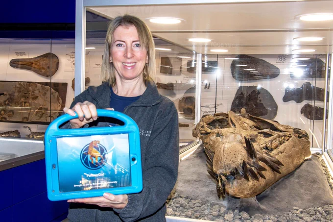 A woman is holding up a tablet and is standing next to a glass cabinet that contains the skull of a Pliosaur