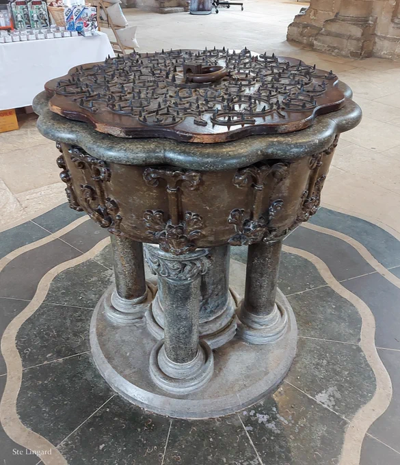 A colour photograph of an ornate font, set on a marble floor. For information about the floor, see:  https://www.stwhitesstone.co.uk/peterborough-cathedral-font/

For more on the financial crisis, see: https://www.peterborough-diocese.org.uk/aboutus/news/cathedralincrisis.php