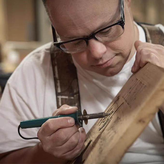 A man wearing glasses focuses intently as he uses a wood-burning tool to create intricate designs on a wooden board, showcasing craftsmanship and attention to detail.