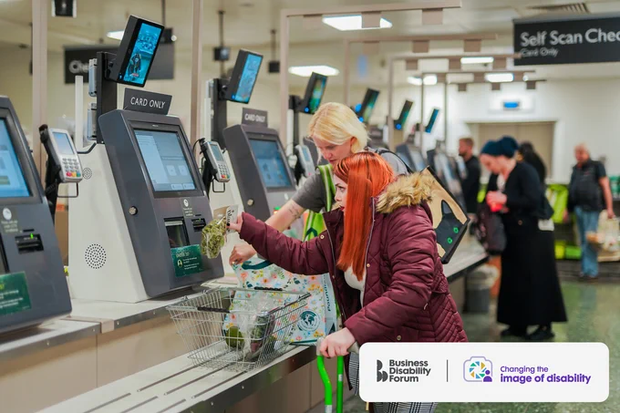 Person with cerebral palsy, who uses bright green walking sticks, scans a product at self checkout in a supermarket while her friend bags purchases. The Business disability Forum 'Changing the image of disability' logo in the bottom right corner of the image.