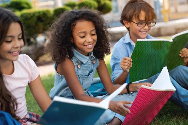 A group of school kids read outside, sitting on the grass.