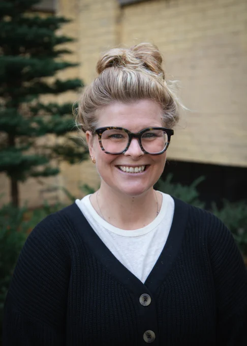 Person with a bun hairstyle and glasses smiling in front of a brick building, wearing a black cardigan over a white top.