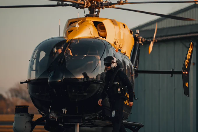 An observer walking in front of Police45 while conducting pre-flight checks. The observer has his visor down and the pilot is working in the front getting the helicopter ready for take off.