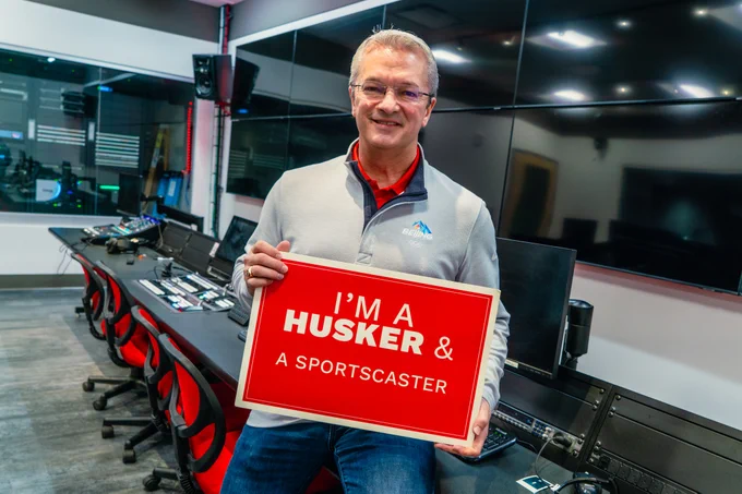 Bill smiles for a photo holding a sign that says "I'm a Husker & a sportscaster"