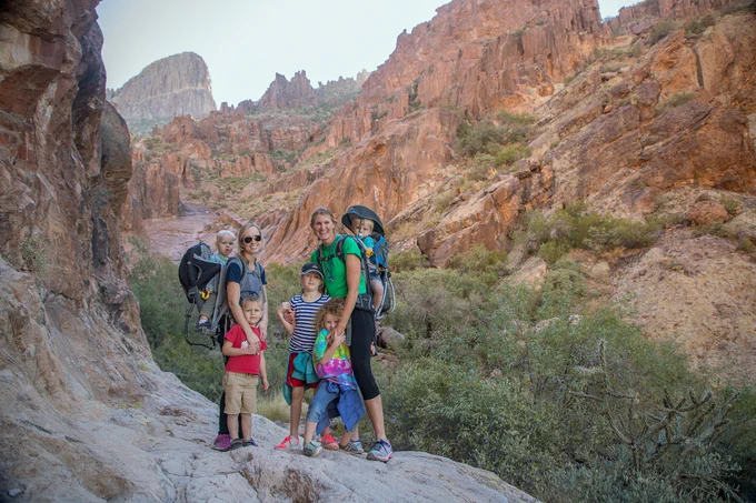 A group of five people with hiking gear, including two carrying young children in backpack carriers, standing on a rocky trail in a canyon with steep, red rocky cliffs in the background.