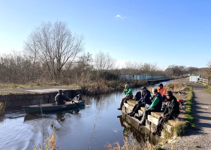 Group of volunteers sit on the bank of the canal in the sunshine taking refreshment while watching a canoe pass by on the glimmering canal water. It looks cold, but the sun shining and the sky is almost cloudless. The trees in the background have lost their leaves.