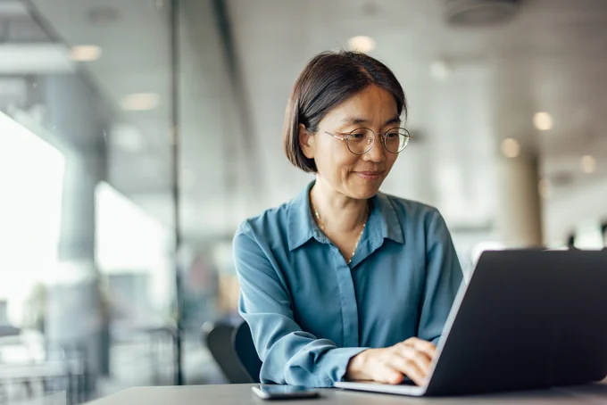 Confident, older Asian woman wearing glasses, reading the news on laptop screen.