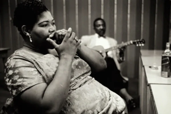 Big Mama Thornton, tocando la armónica en un camerino. Es una mujer afroamericana voluminosa que lleva un vestido ceñido a flores y tiene anillos en las manos. Al fondo, desenfocado, un guitarrista también afroamericano con pajarita y chaqueta descansa con una guitarra en las manos. A la derecha, sobre una mesa, destaca una botella de whisky o bourbon. Dominio público.
