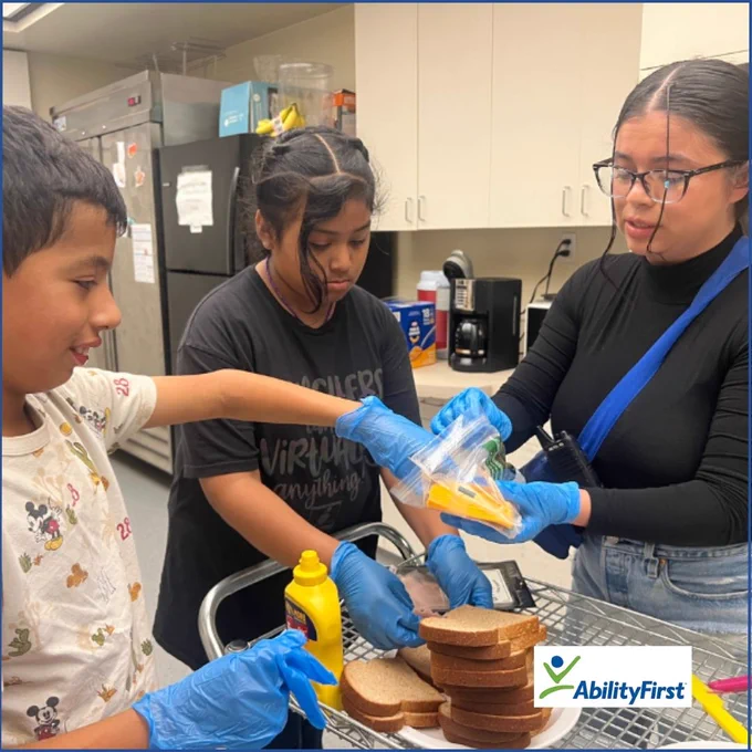 Three individuals stand in a kitchen making sandwiches wearing blue gloves. They add mustard to the sandwiches. The two girls wear black and the boy wears a white mickey mouse shirt. The AbilityFirst logo is in the bottom-right corner. 

