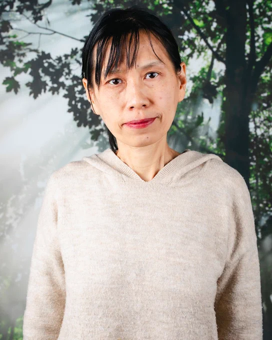 Asian woman with black hair wearing beige sweater, standing in front of a picture of a tree with sunlight streaming through the branches