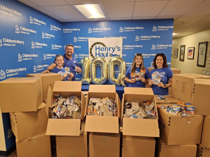 Five volunteers at Children's Mercy Kansas City posing with boxes of donations in a room decorated with "Henry's Haul" posters. They are wearing matching blue t-shirts and are standing behind large cardboard boxes filled with various items.