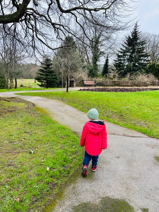 A small child wearing a green woolly hat and a red coat walks through a park.