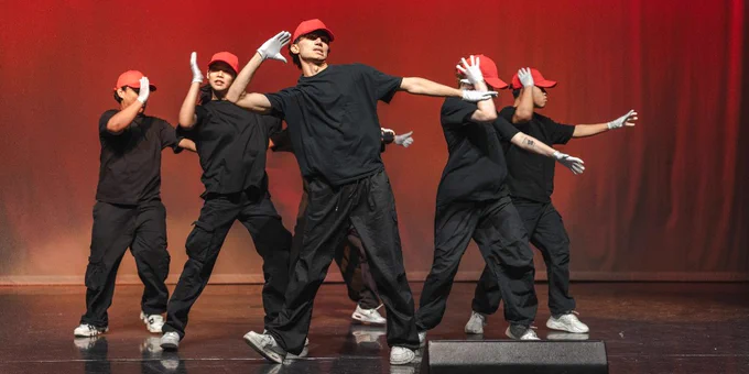 A group of urban dancers in all black clothing, white trainers, and red caps preforming on large stage with red backdrop.