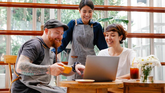 Three people in a café setting, two baristas and one customer, smiling and collaborating over a laptop with coffee and iced drinks on the table.