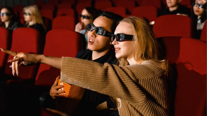 A young man and woman wearing 3D glasses, enjoying a movie in a theater with popcorn, surrounded by other viewers.