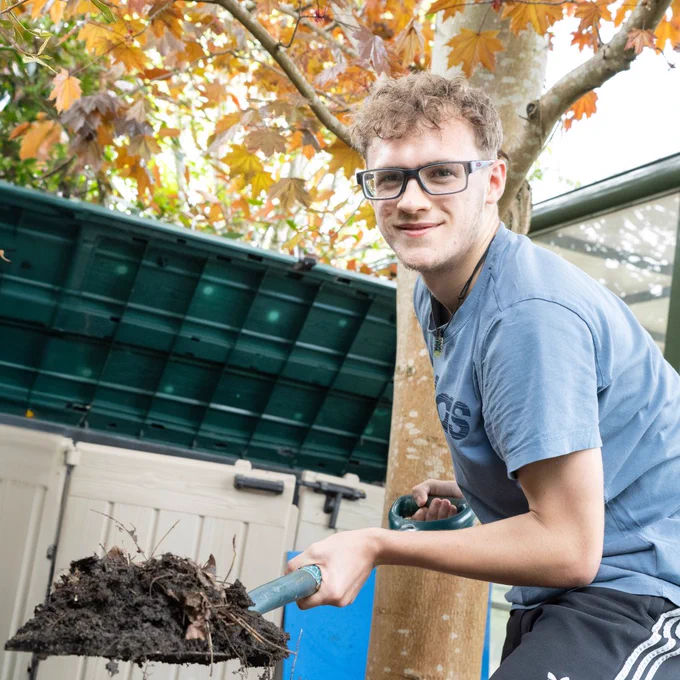 An Inspire learner is holding a shovel of mud whilst smiling at the camera.