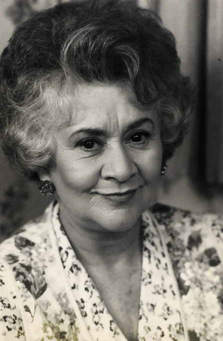 A black and white image of Dame Joan Plowright. They are wearing a white floral blouse, with short curled hair and earrings.