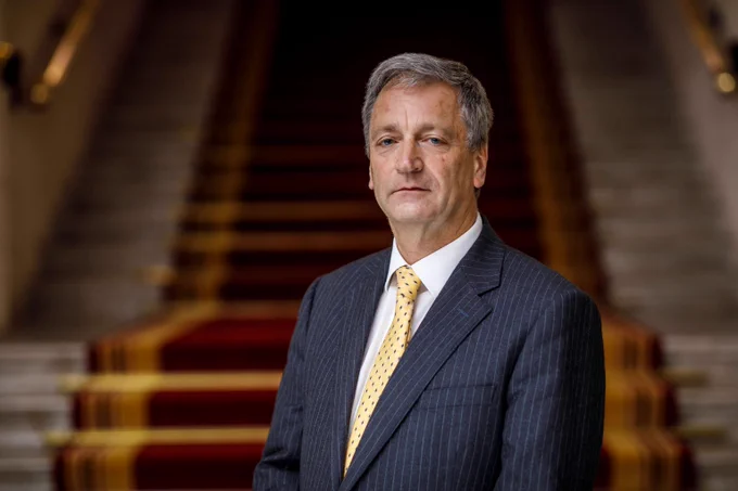 Photo of Richard Atkinson. White man with grey hair wearing a black striped suit, yellow tie and white shirt standing by some stairs in the Law Society.