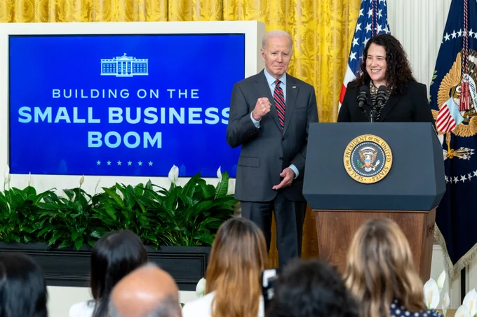 President Joe Biden stands next to a person at a podium in the White House, addressing an audience under a sign reading "Building on the Small Business Boom.