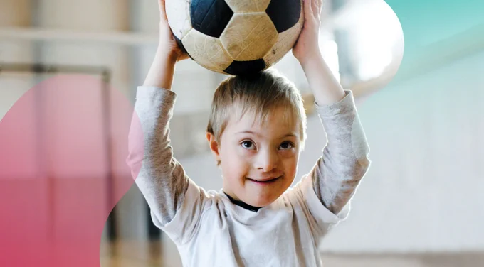 white, blonde-haired boy with Down syndrome wearing white t-shirt over grey long-sleeved shirt holds a soccer ball on his head in a gymnasium
