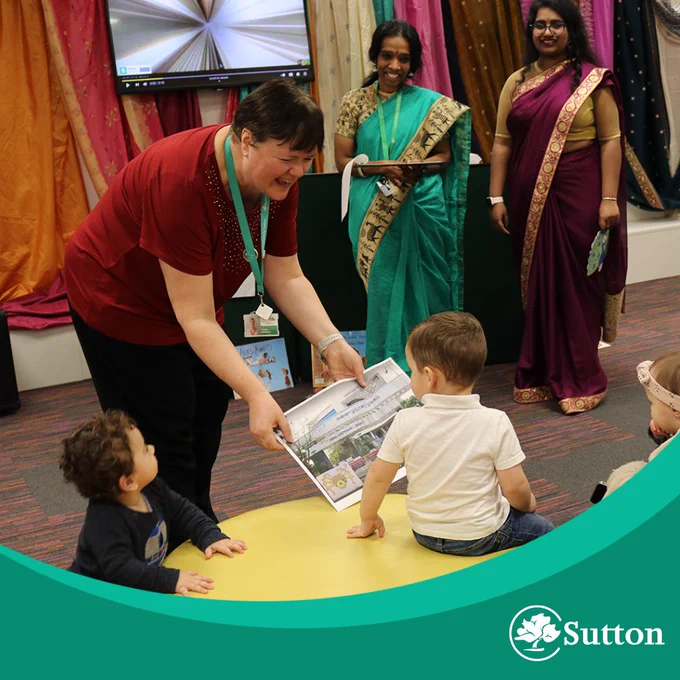 Two women in traditional Indian saris are standing in front of a colorful display of fabric.The woman on the left wears a teal saree with gold accents, while the woman on the right wears a dark pink saree with a gold border. They are smiling. In the foreground, a woman with short brown hair showing a book to two young children, who are turned towards the women. The background features a wall covered in fabrics of many different colours.