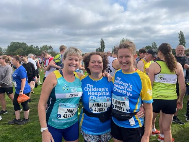 Three women stand in a field with lots of runners. They are smiling at the camera and are dressed for a run. 