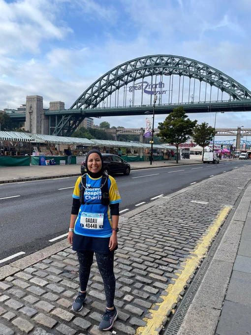 A woman in running gear stands in front of the Tyne bridge in Newcastle. The bridge has a sign on in that reads "Great North Run". 