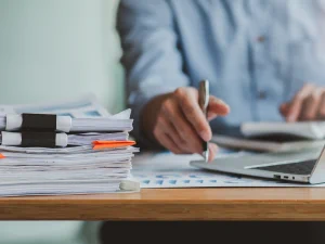 A person sitting at a desk with a laptop and a stack of papers in binder clips.