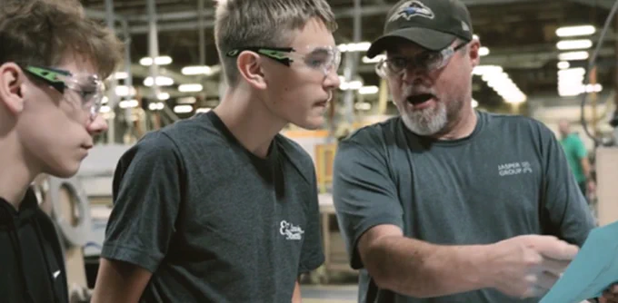 Two high school students learn from a technical education instructor in a large warehouse.