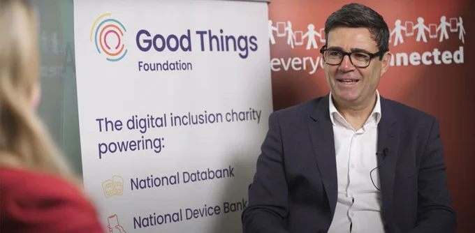 Mayor of Greater Manchester Andy Burnham sitting opposite Helen Milner OBE (Group CEO of Good things Foundation). Andy has glasses and is smiling. He is sitting next to a banner with Good Things' logo on it and the text reads 'The digital inclusion charity powering: National Device Bank and National Device Bank'