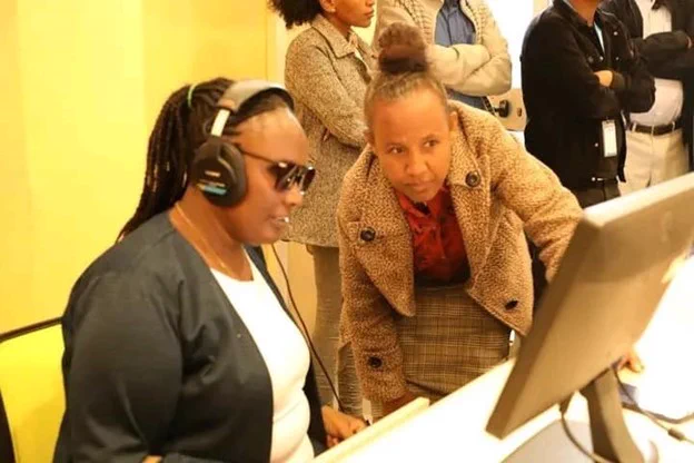 A participant with a visual impairment at the Dessie Civic Incubation Center hosted by Wollo University. Two individuals are observing a computer screen, one wearing headphones and operating the computer, while the other looks on attentively. They are in a room with other people in the background. 