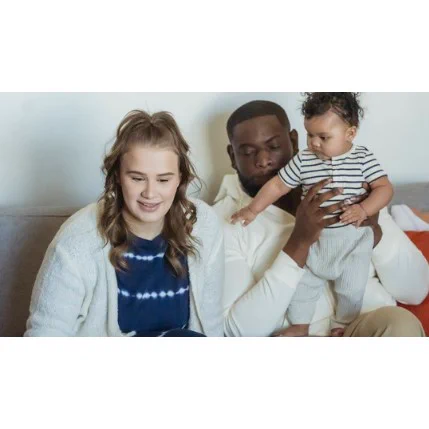 A young family sitting on a couch. A woman with wavy light brown hair and a white cardigan smiles softly while looking forward. Beside her, a man with short dark hair and a white shirt gently holds a baby. The baby, wearing a striped onesie and light gray trousers, reaches out curiously.