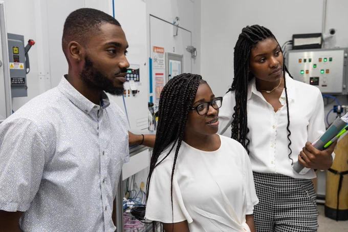 Three black HBCU students wear business casual attire. The trio stands together listening to a speaker during a laboratory tour. 

Credit: NASA