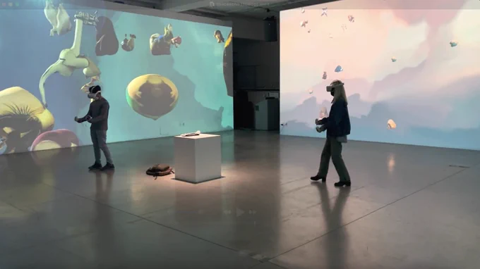 In a large room in an art gallery, two visitors in the center wear VR headsets. Two gallery walls display 3D video game renderings of pastel-colored shapes and clouds. 