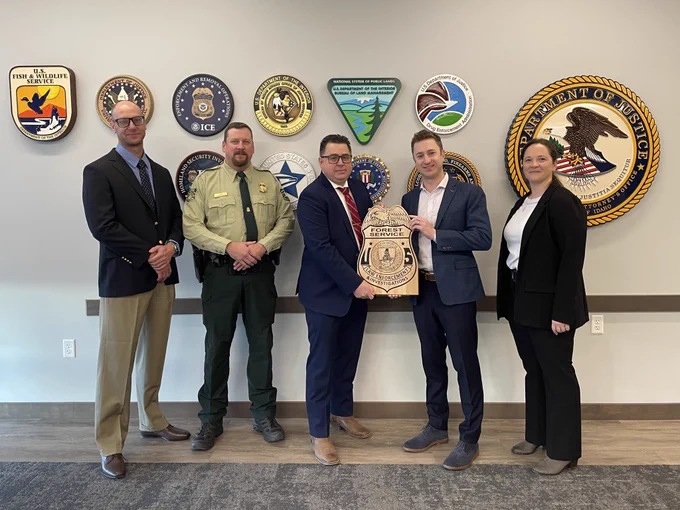 U.S. Attorney Josh Hurwit and four representatives from the United States Forest Service standing in front of several agency seals and holding the USFS wooden seal.