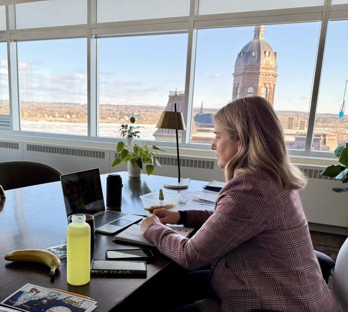 Premier Susan Holt sits at a desk in an office, focused on a laptop. Papers, a banana, a water bottle, and a notebook are on the table. Large windows reveal a cityscape with the New Brunswick Legislature. Lush plants adorn the space.